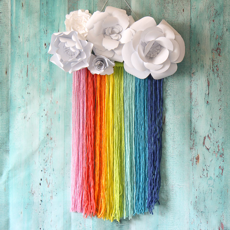 This easy rainbow craft features a rainbow yarn macrame wall hanging topped with paper flower clouds.
